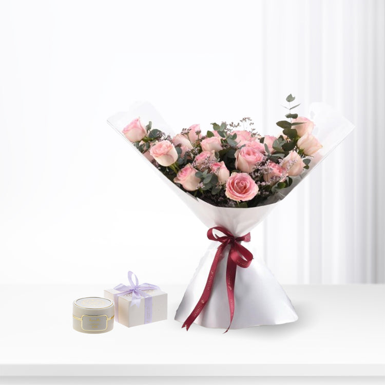Soft pink roses in white paper, tied with burgundy ribbon, beside elegant gift boxes.