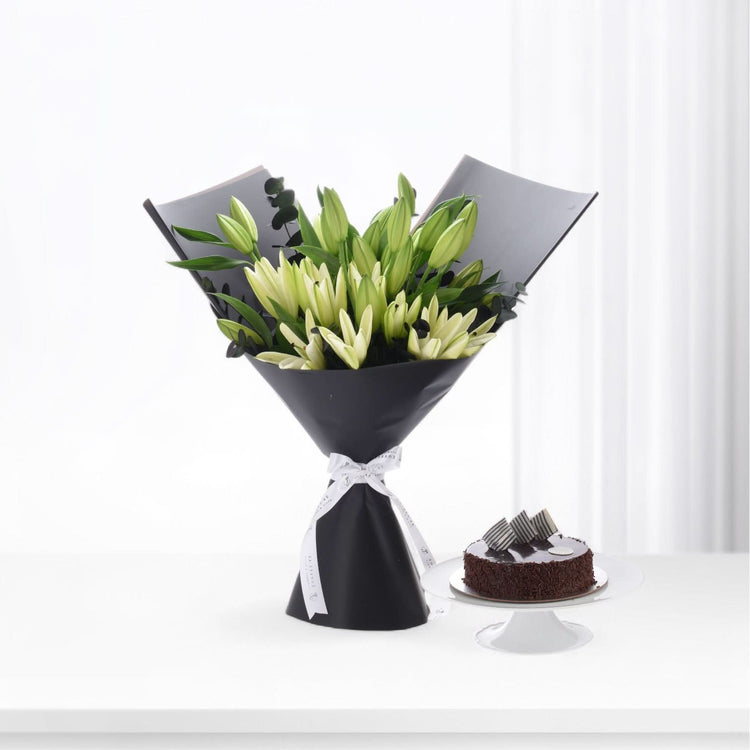 Elegant black-wrapped white lily bouquet beside chocolate cake on white pedestal.