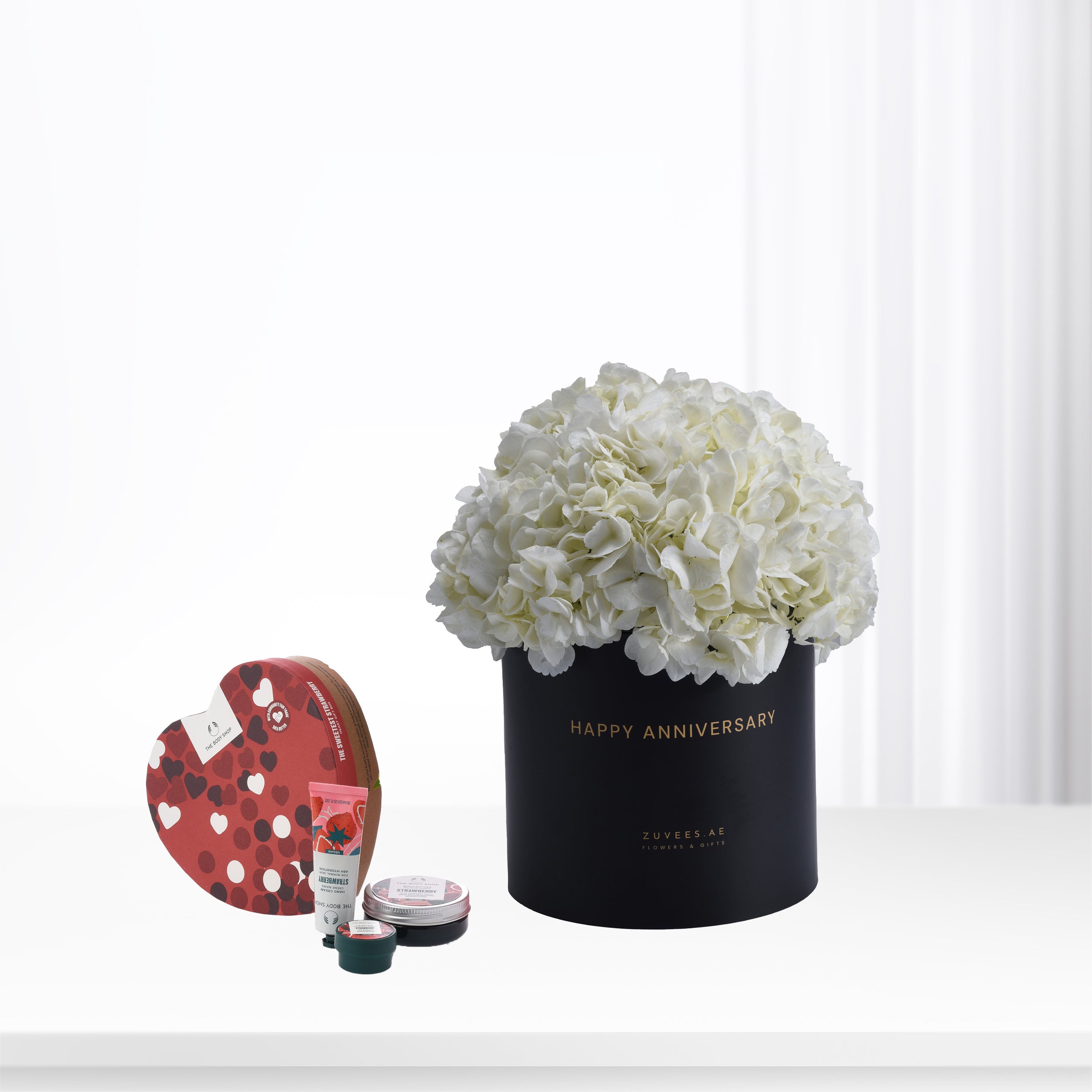 Elegant white hydrangeas in black "Happy Anniversary" box with heart-themed skincare set.