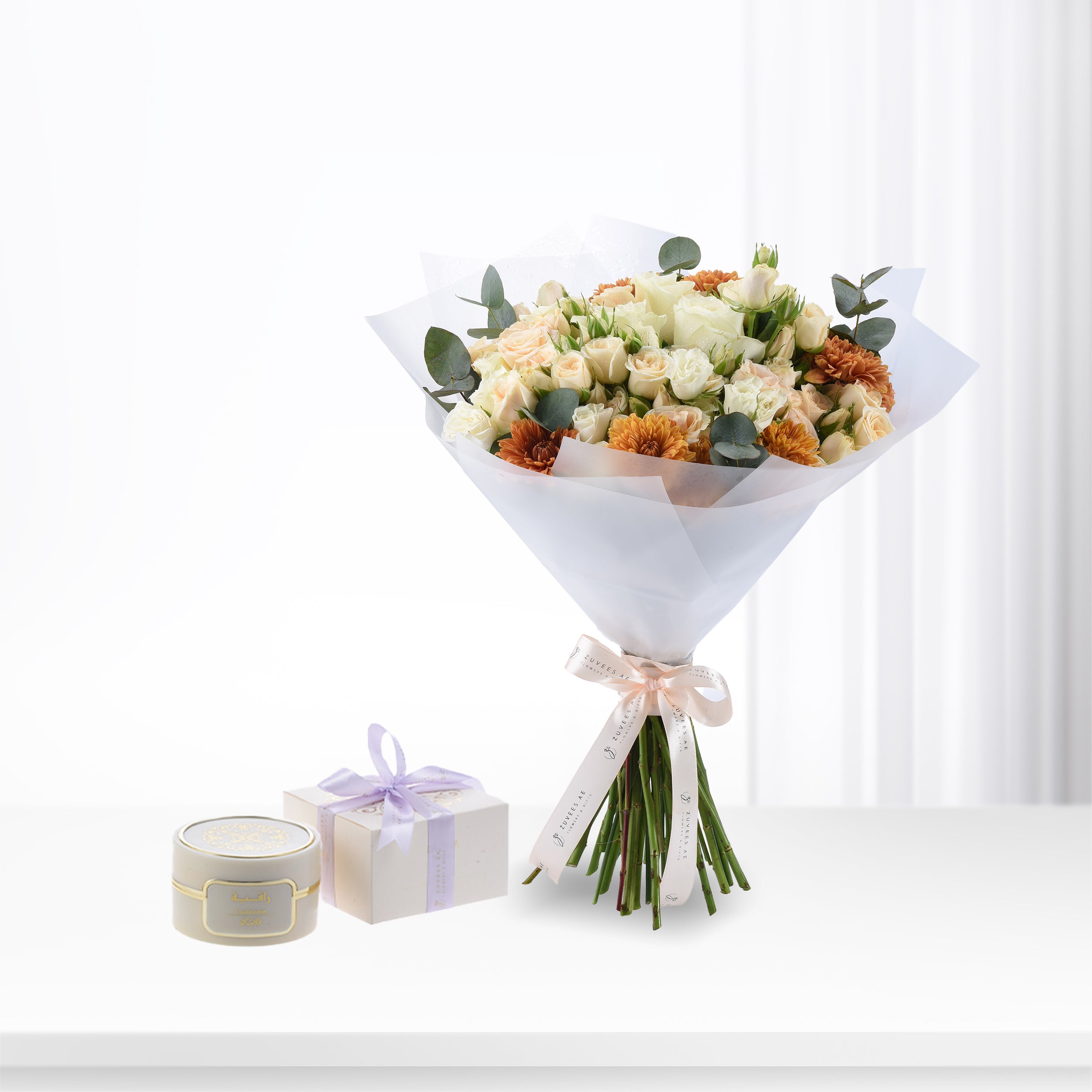 Soft peach roses and orange dahlias wrapped in white paper with branded ribbon, beside elegant gift box and candle.