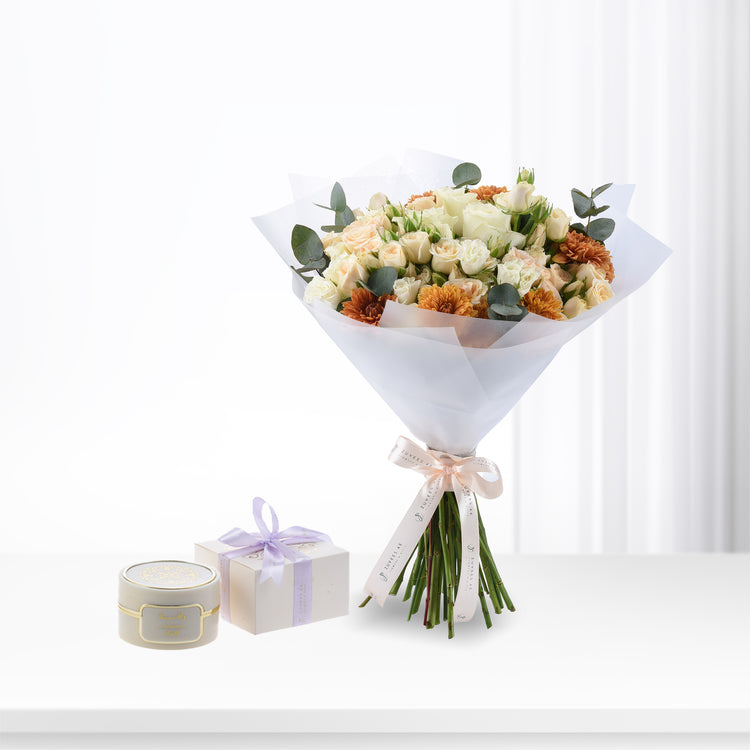 Soft peach roses and orange dahlias wrapped in white paper with branded ribbon, beside elegant gift box and candle.