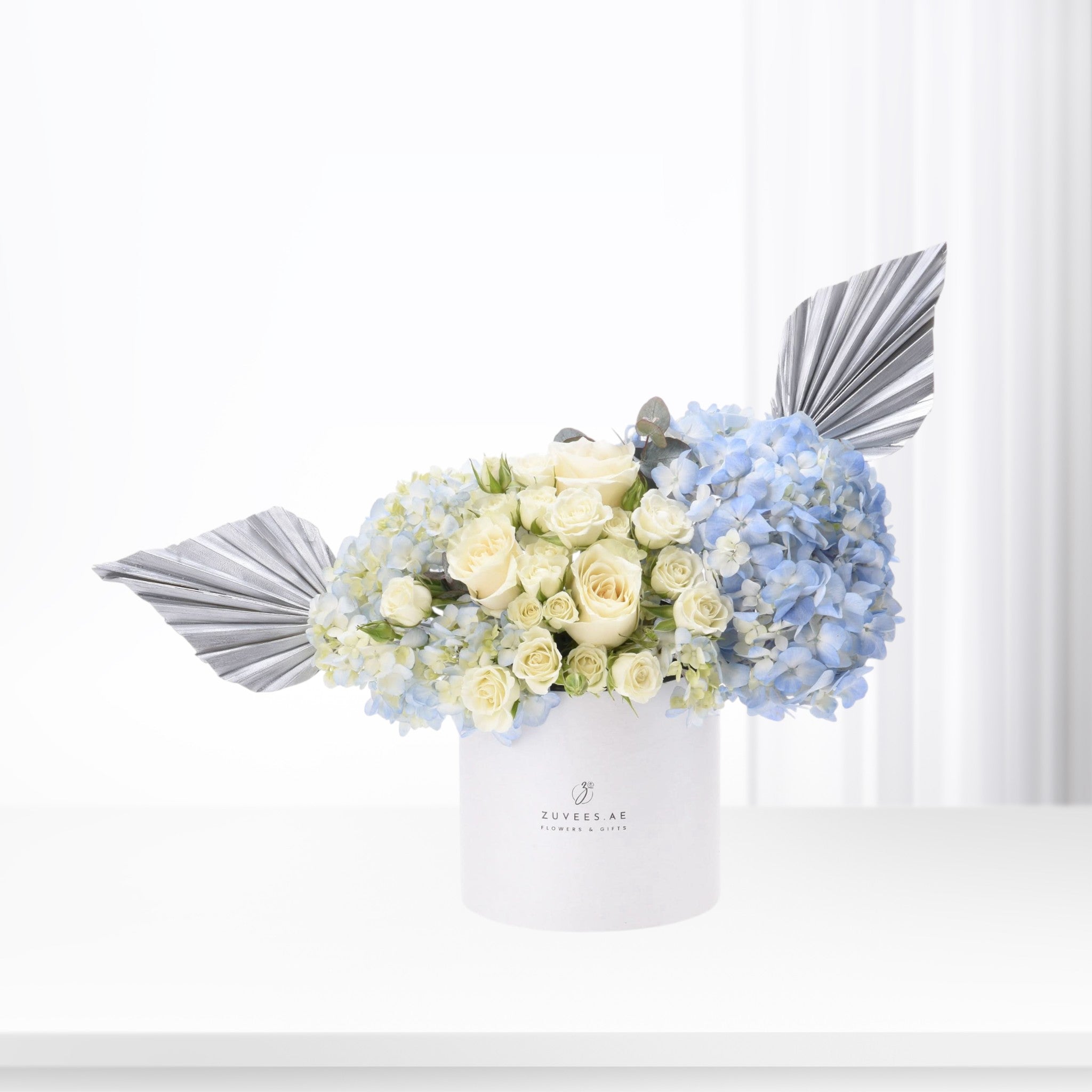 Elegant white roses and blue hydrangeas in white box with silver fan accents, minimalist luxury design.