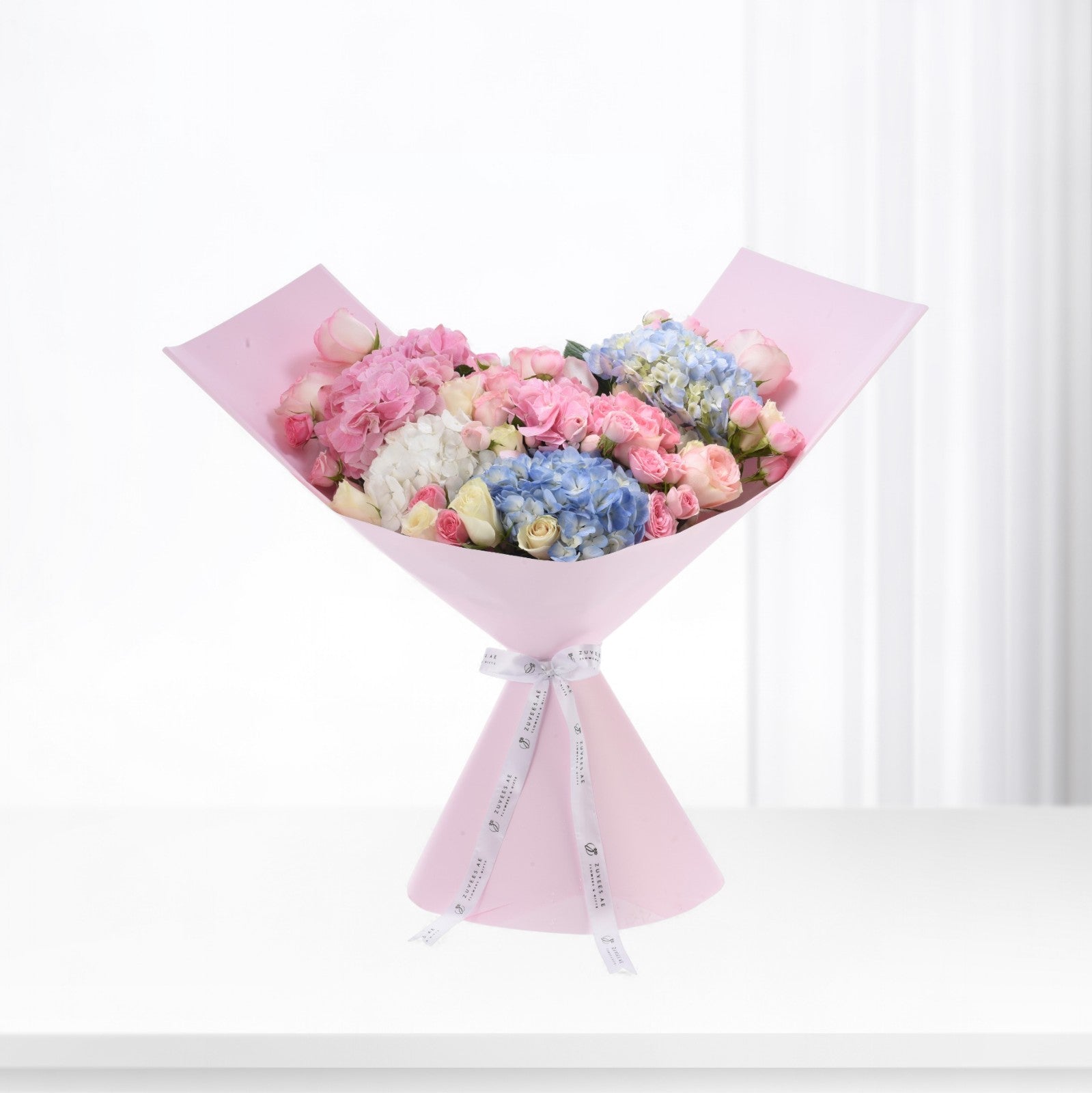Pastel pink, blue, and white hydrangeas and roses elegantly wrapped in soft pink paper with branded ribbon.
