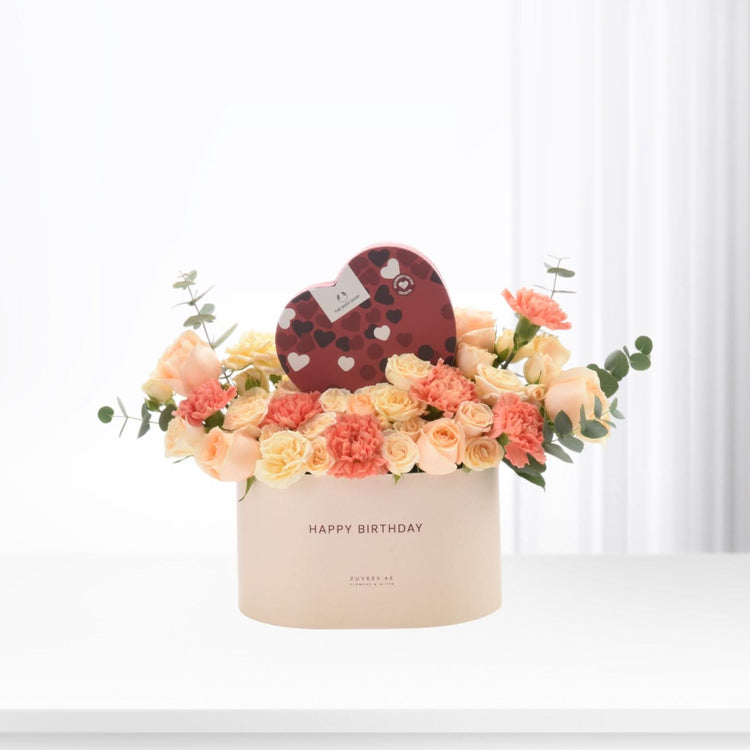 Pastel peach roses and carnations in cream box with heart chocolate gift.