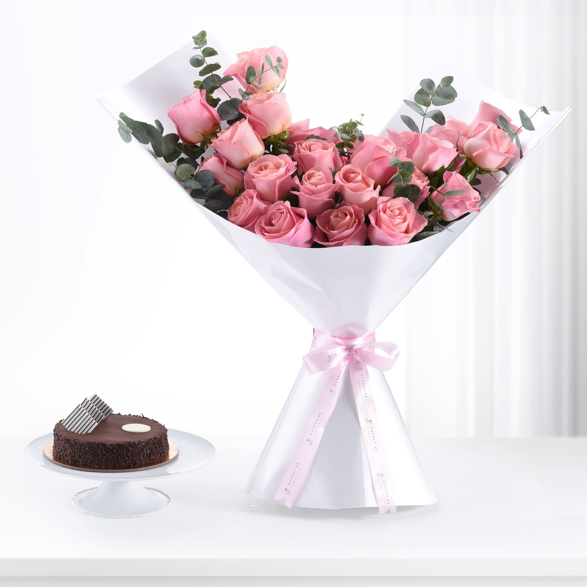 Pink roses in white wrap with pink ribbon, beside chocolate cake on white stand.