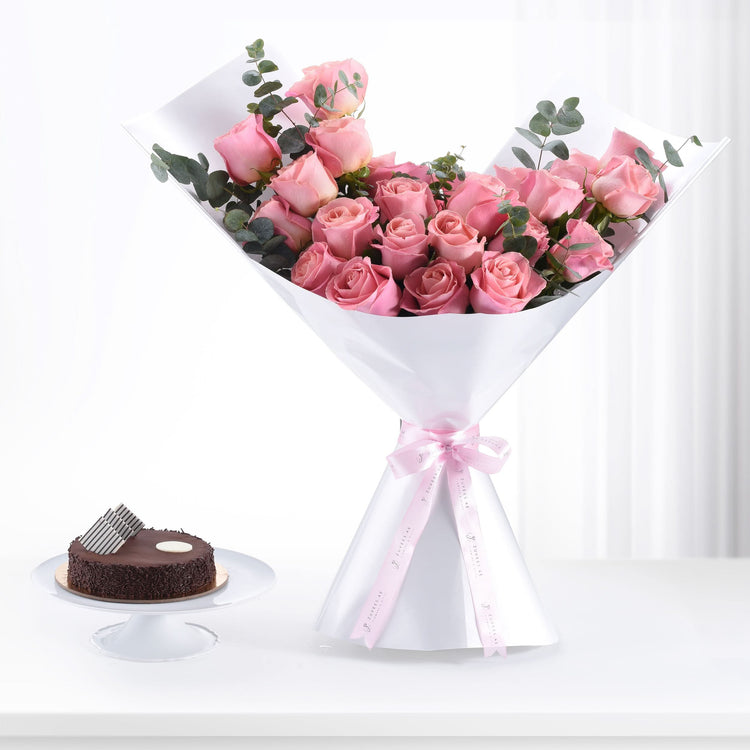 Pink roses in white wrap with pink ribbon, beside chocolate cake on white stand.