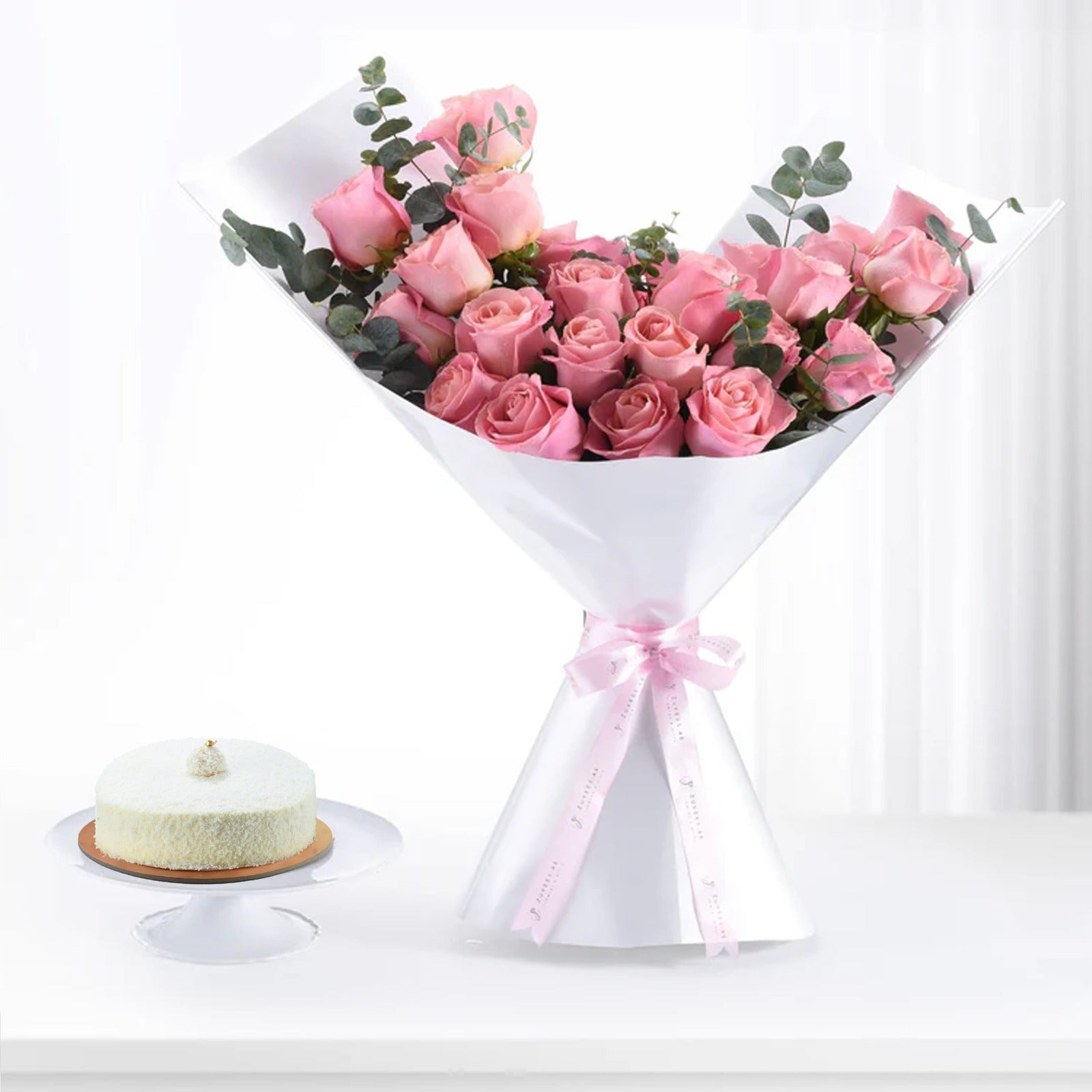 Soft pink roses in white paper, tied with ribbon, beside a delicate cake.