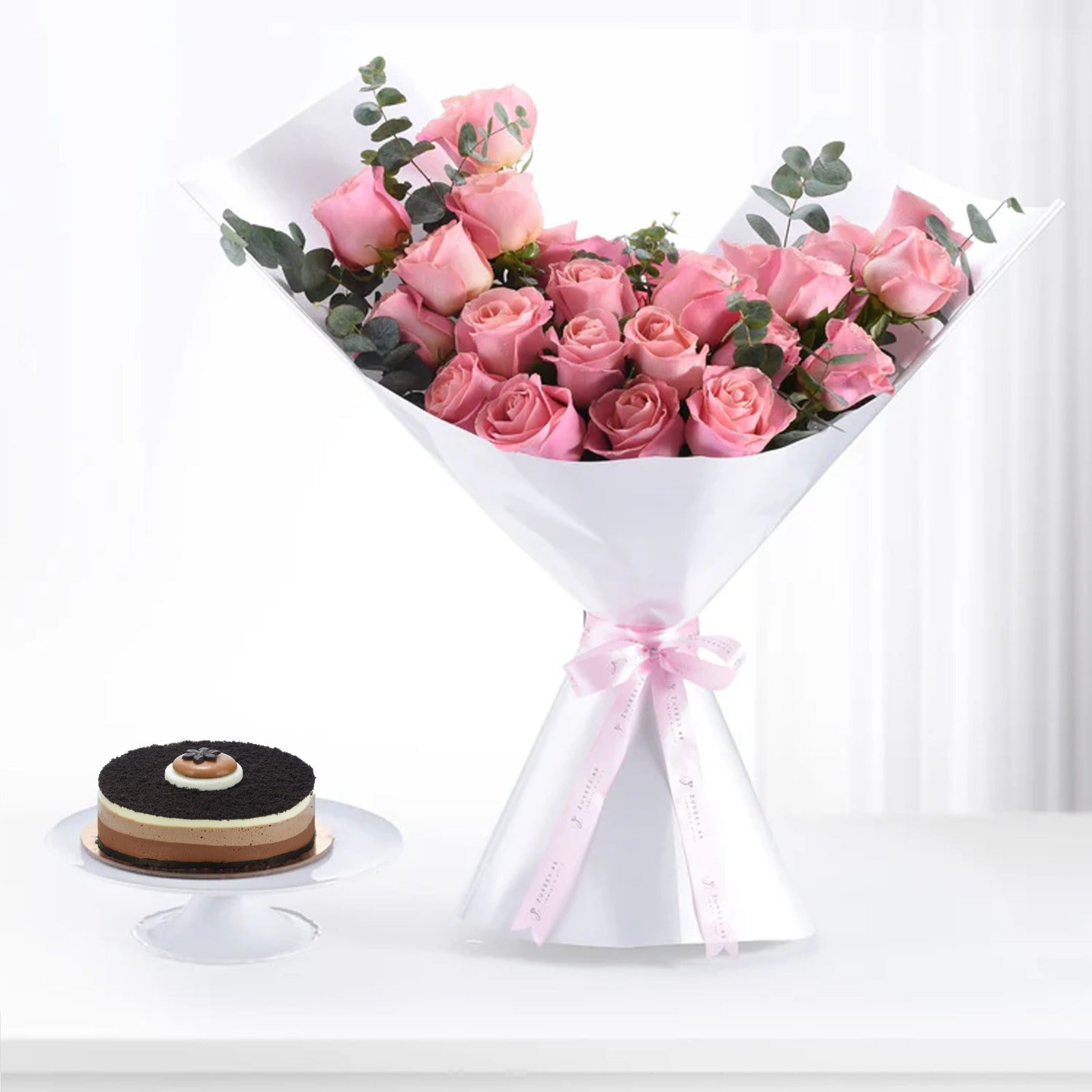 Pink roses bouquet with eucalyptus, white wrap, pink ribbon, beside chocolate cake.