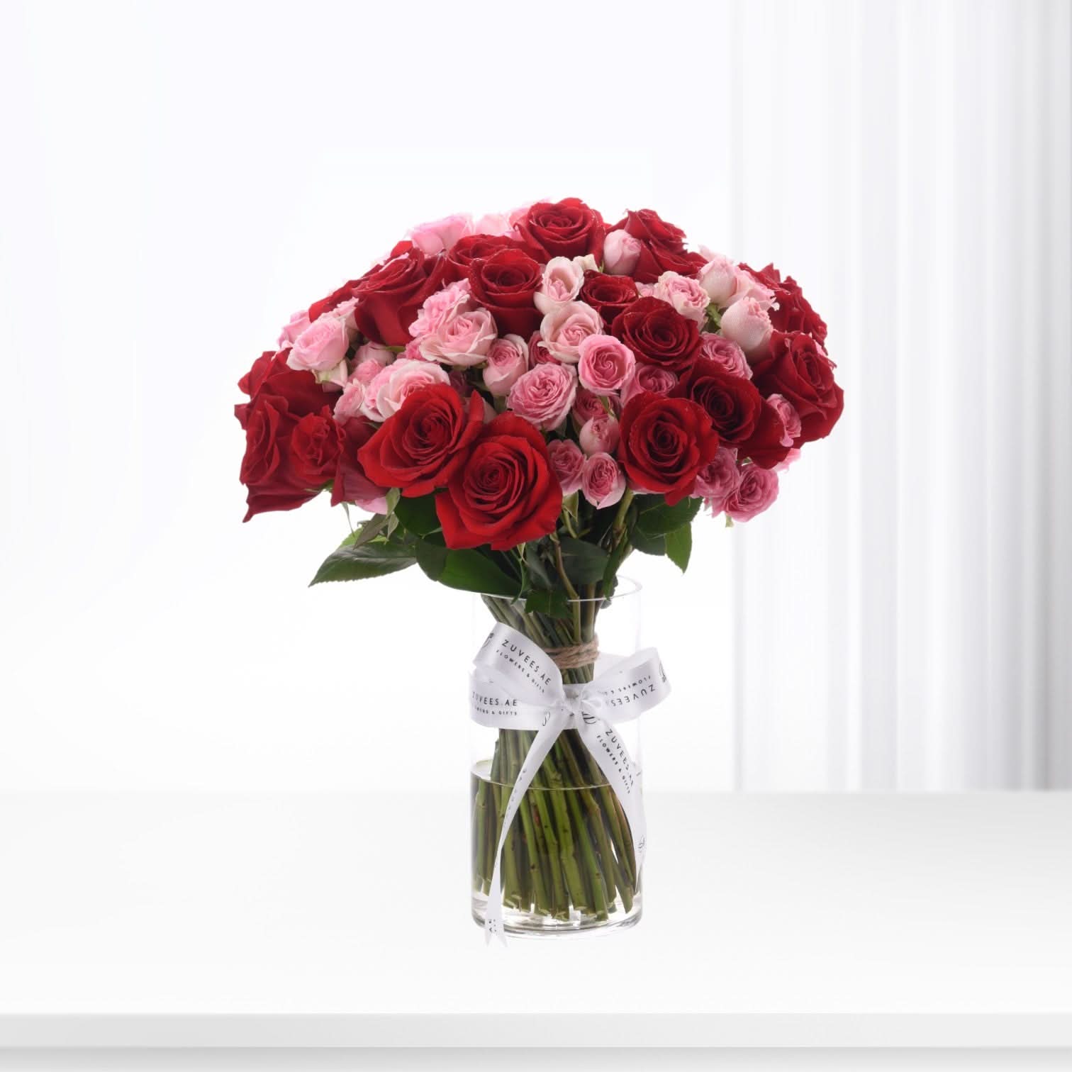 Vibrant red and pink roses in clear vase, elegantly tied with branded white ribbon.
