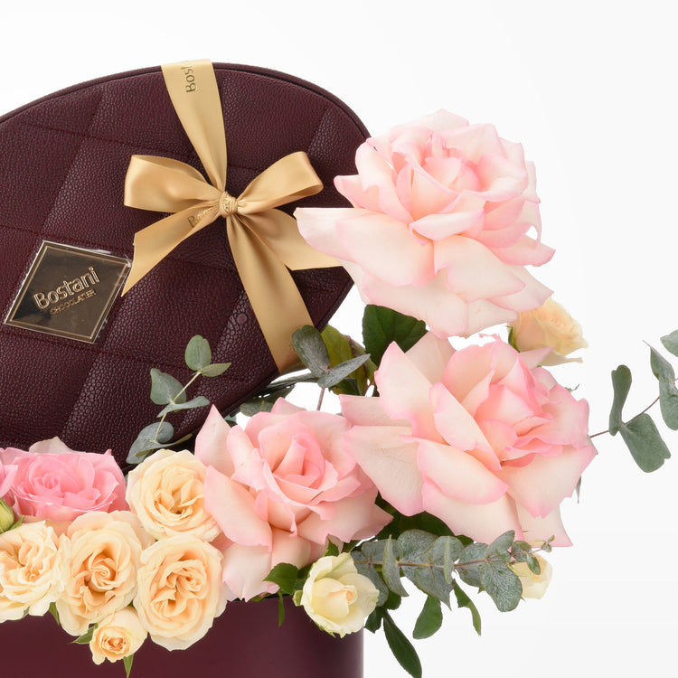 Luxurious burgundy chocolate box with gold bow, nestled among soft pink and cream roses.