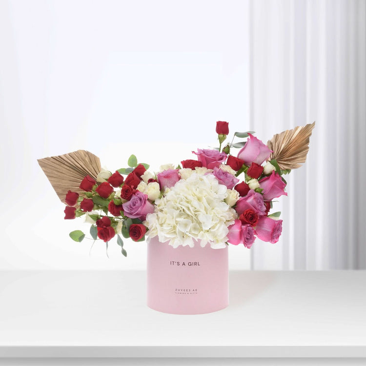 Pink box holds vibrant red, pink, and white blooms with gold accents.
