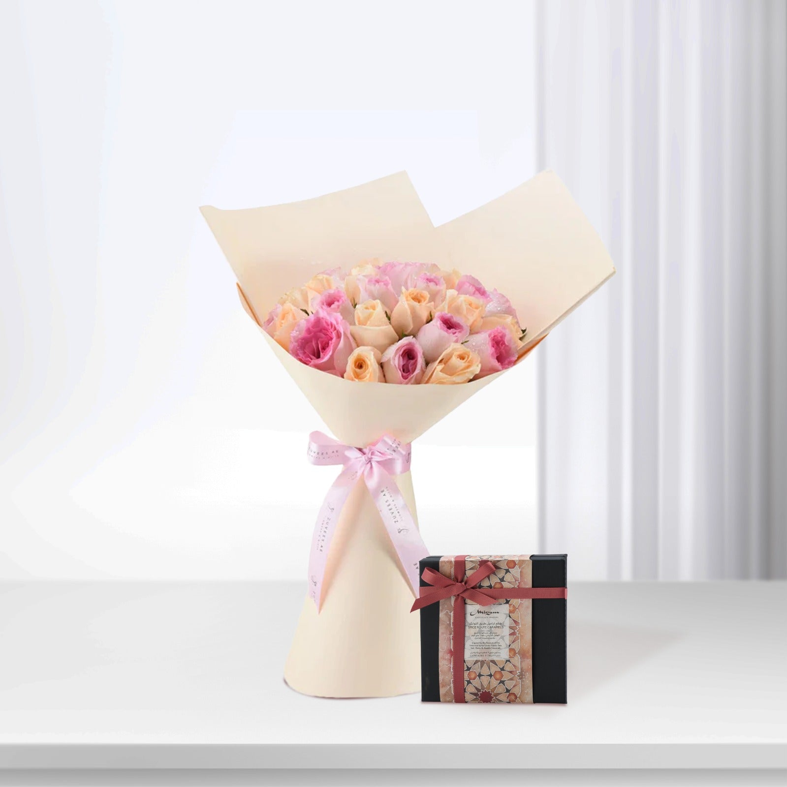 Soft pink and peach roses wrapped in cream paper with pink ribbon, beside patterned chocolate box.
