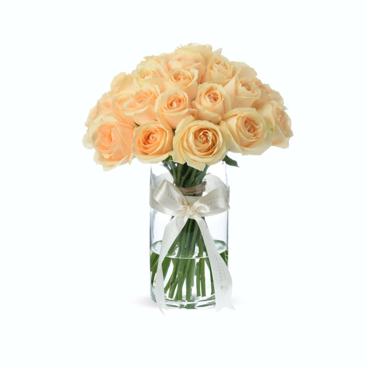 Soft peach roses in clear glass vase, elegantly tied with cream ribbon.