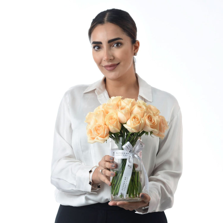 Woman holds peach roses in branded clear vase against white background.