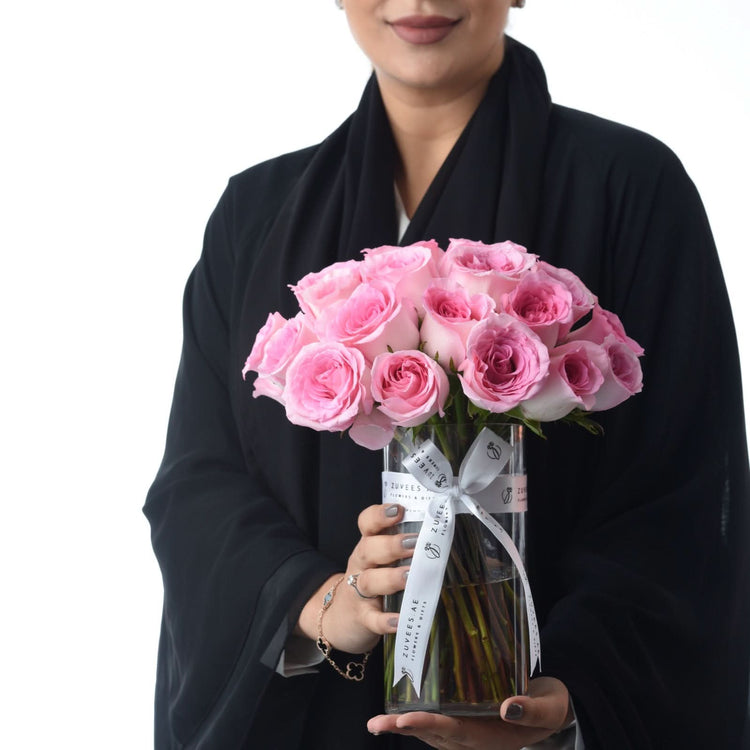 Pink roses in clear vase, white ribbon, held by woman in black abaya, elegant gift presentation.