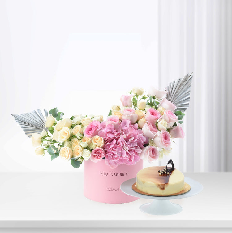 Pink floral arrangement with angel wings, beside cheesecake on stand.