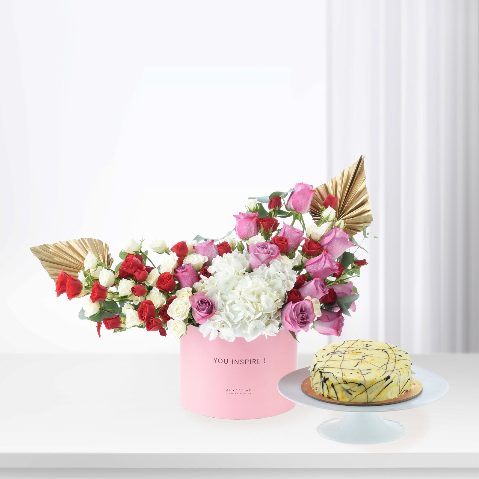 Pink box holds vibrant red, pink, white roses and hydrangeas, accented with gold leaves. Beside, yellow cake with chocolate drizzle.