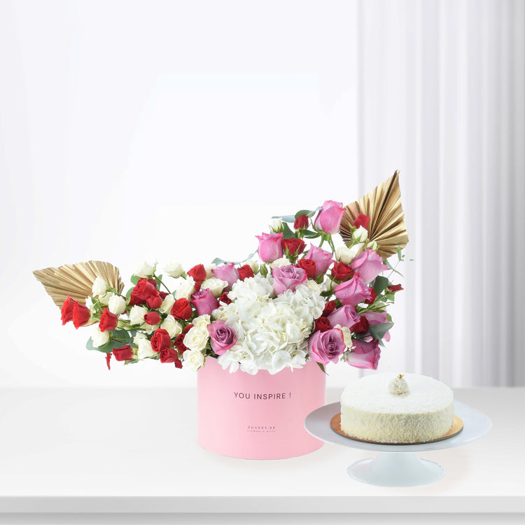 Pink box holds vibrant red, pink, and white roses with hydrangeas, accented by gold fan leaves, beside a coconut cake.