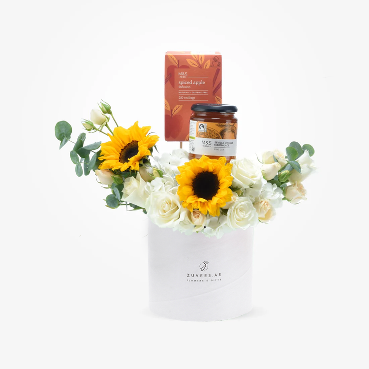 White cylindrical box holds vibrant sunflowers, white roses, and greenery, topped with M&S tea and marmalade.