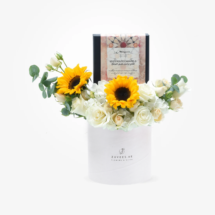 White cylindrical vase holds vibrant sunflowers and cream roses, topped with spice caramel gift box.