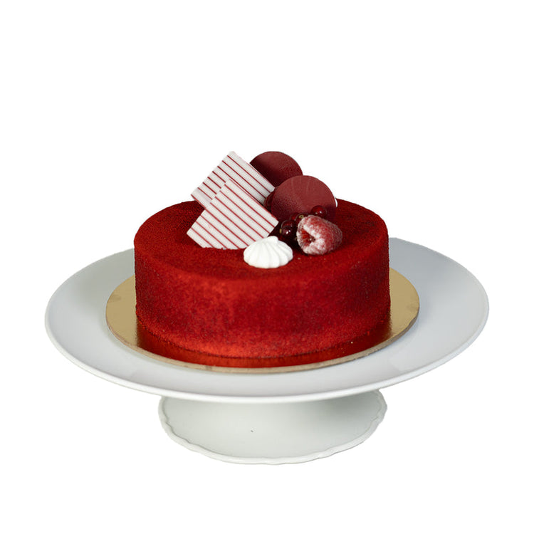 Vibrant red velvet cake adorned with berries, striped chocolate, and cream on elegant white stand.