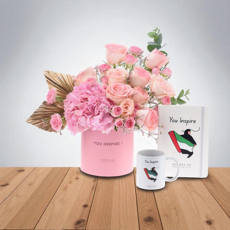 Pink roses and hydrangeas in branded pink vase, matching mug and notebook on wooden table.
