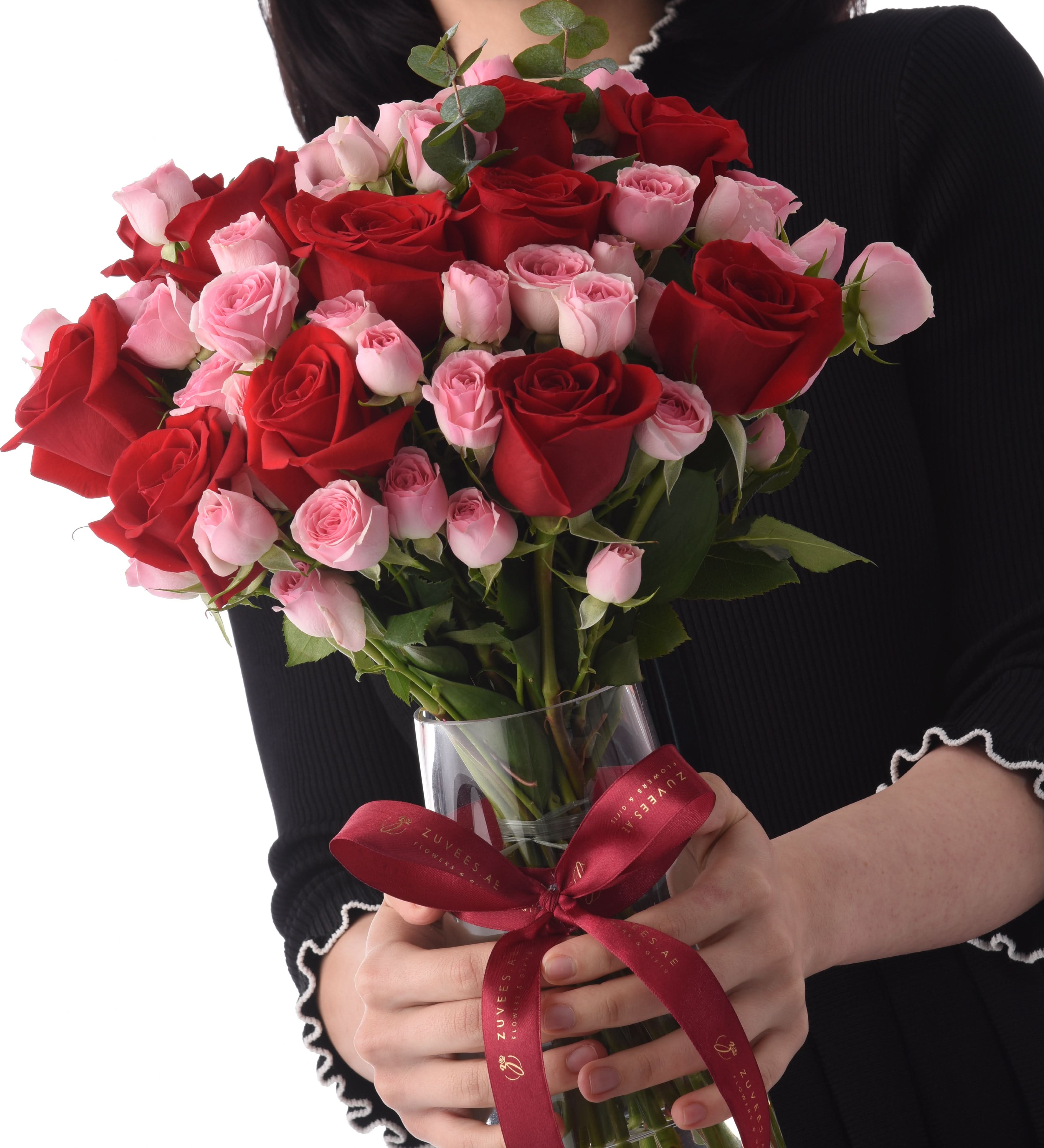 Vibrant red and pink roses in clear vase, tied with branded burgundy ribbon, held by person in black.