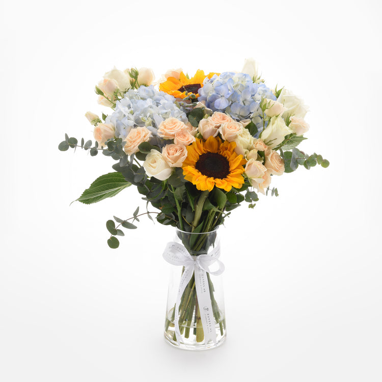 Sunflowers, hydrangeas, and roses in clear vase with white ribbon.