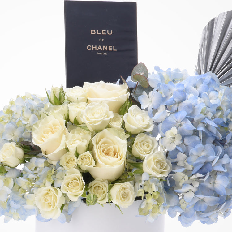 Cream roses and blue hydrangeas surround Chanel Bleu perfume box.