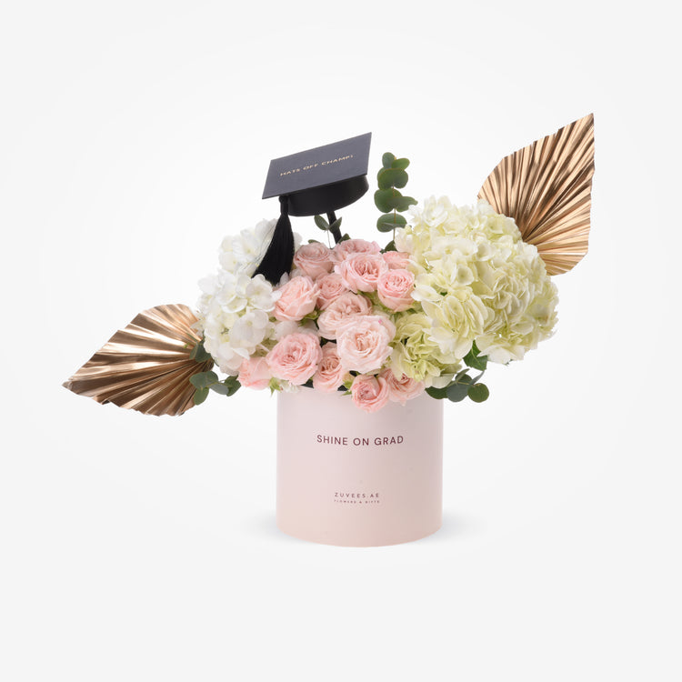 Pink cylinder holds soft roses, hydrangeas, gold leaves, graduation cap.