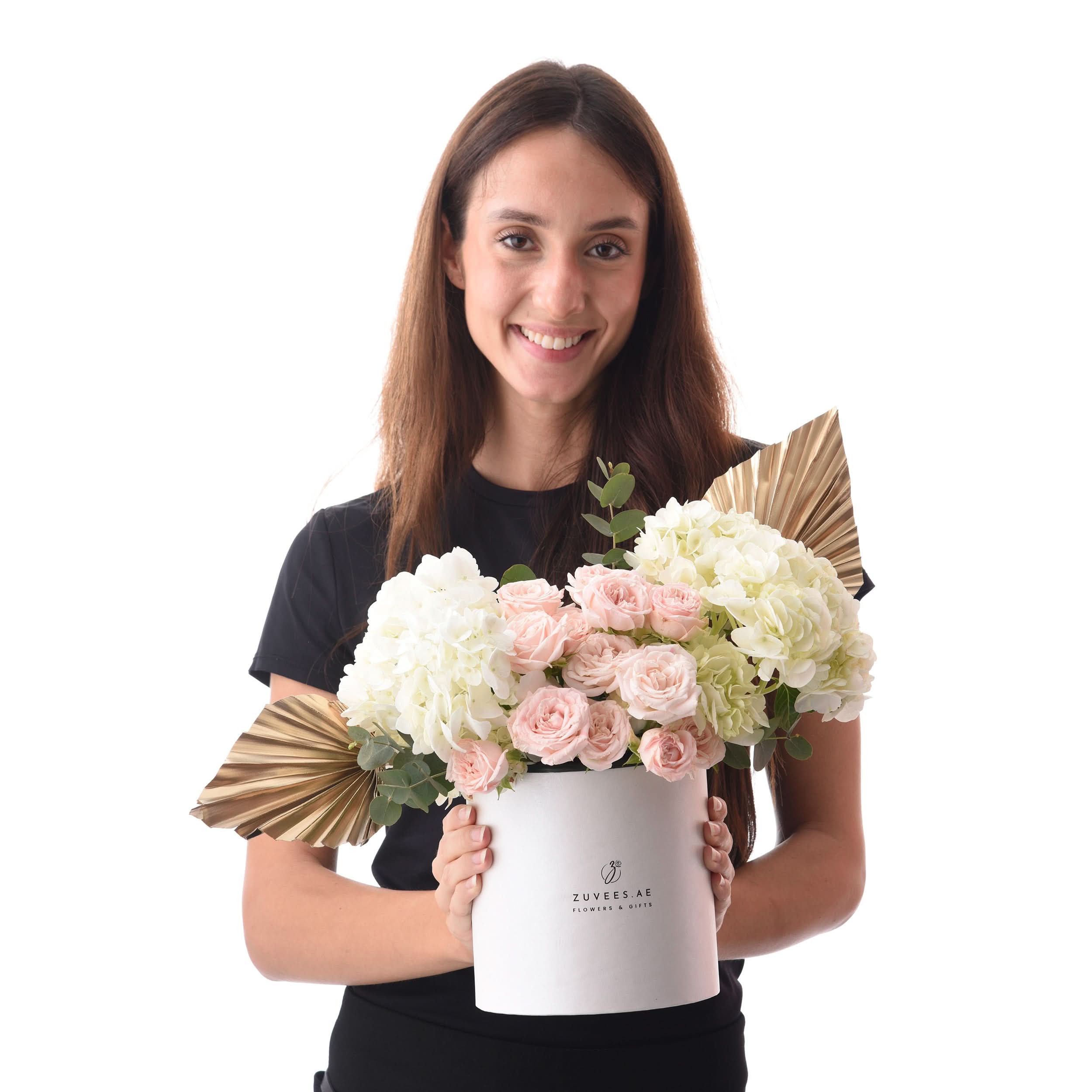 Woman holds elegant white box filled with pink roses and hydrangeas, accented with gold fan leaves.