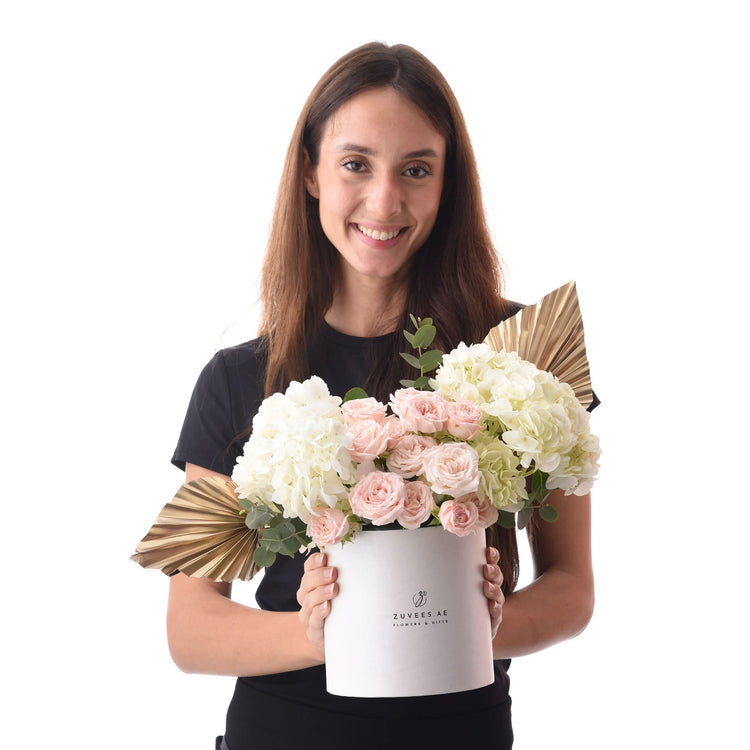 Woman holds elegant white box filled with pink roses and hydrangeas, accented with gold fan leaves.