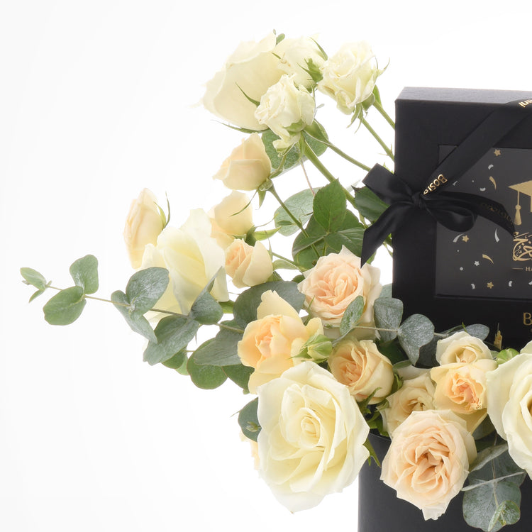 Cream roses and eucalyptus flank a black graduation gift box with gold accents.