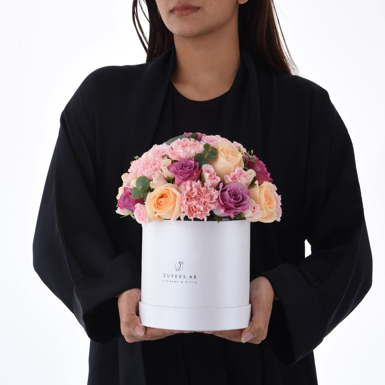 Elegant white box bouquet with pink, peach, purple roses and carnations, held by woman in black.