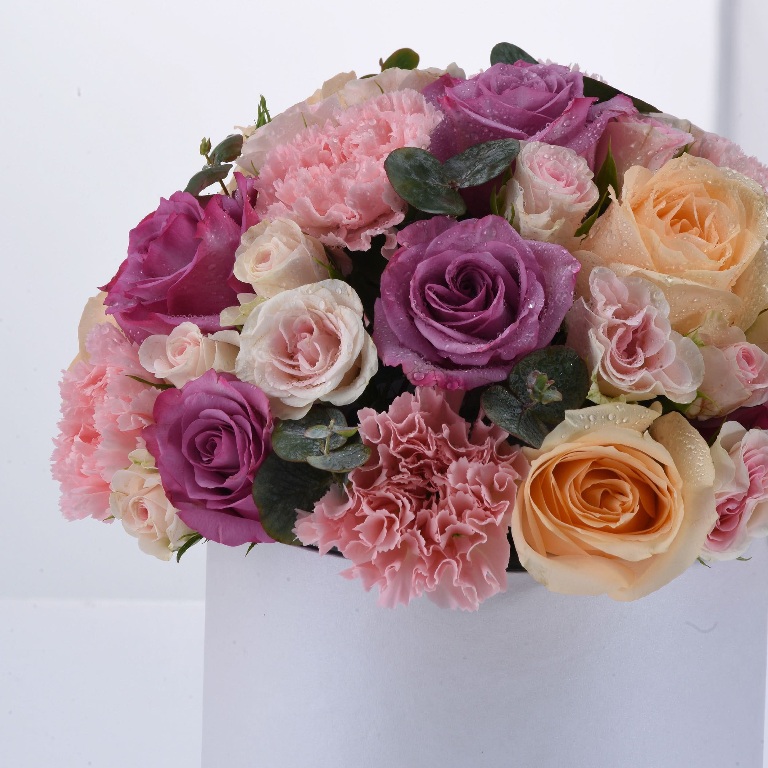 Vibrant pink, purple, and peach roses with carnations, dewdrops, and lush greenery in white box.
