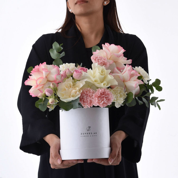 Elegant pink and cream roses with carnations in white Zuvees box, held by woman in black.