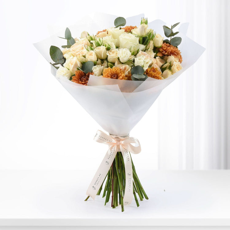 Cream roses, orange chrysanthemums, eucalyptus, wrapped in white paper with branded ribbon.