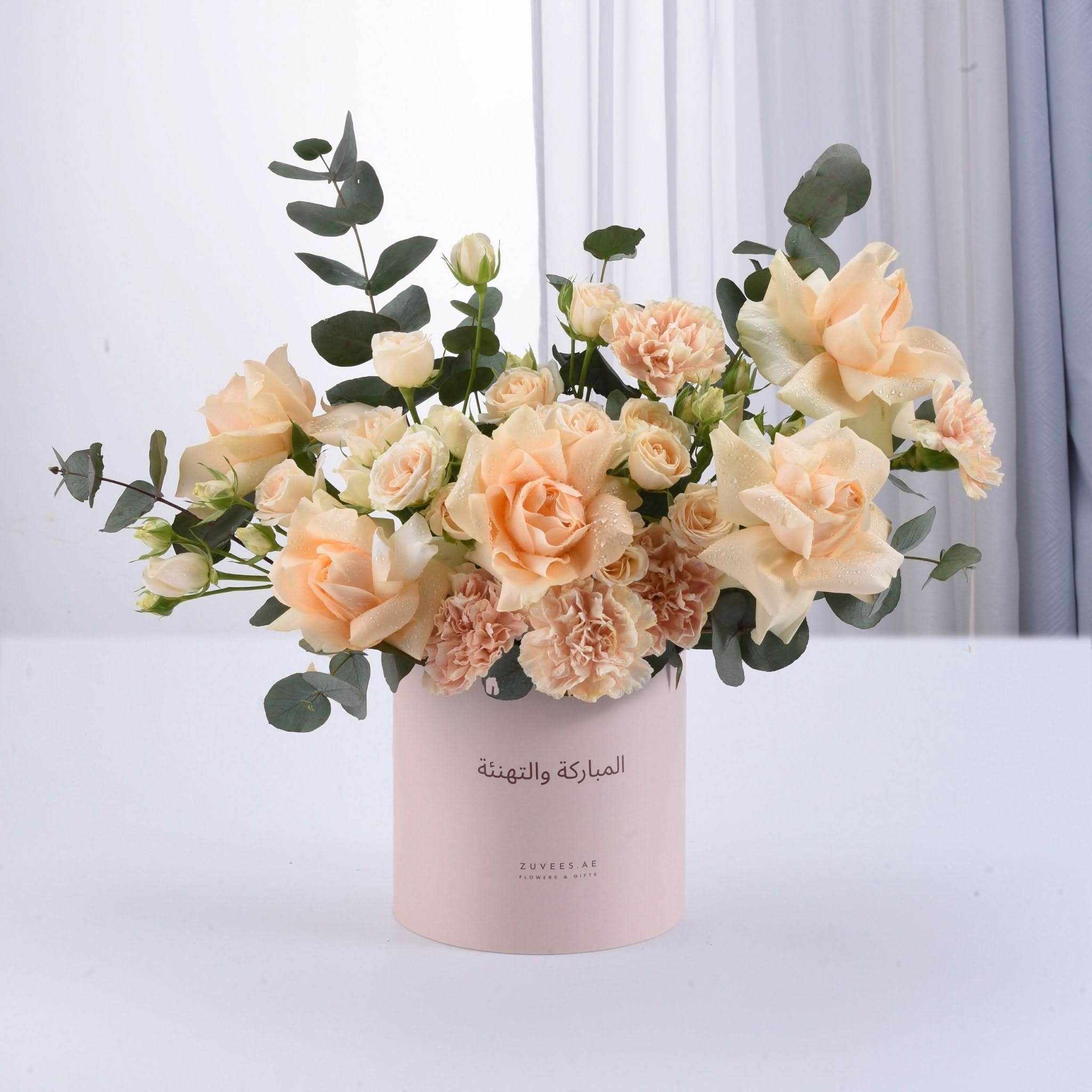 Soft peach roses and carnations in pink cylinder with Arabic greeting.