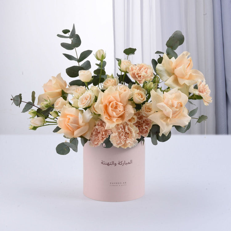 Soft peach roses and carnations in pink cylinder with Arabic greeting.