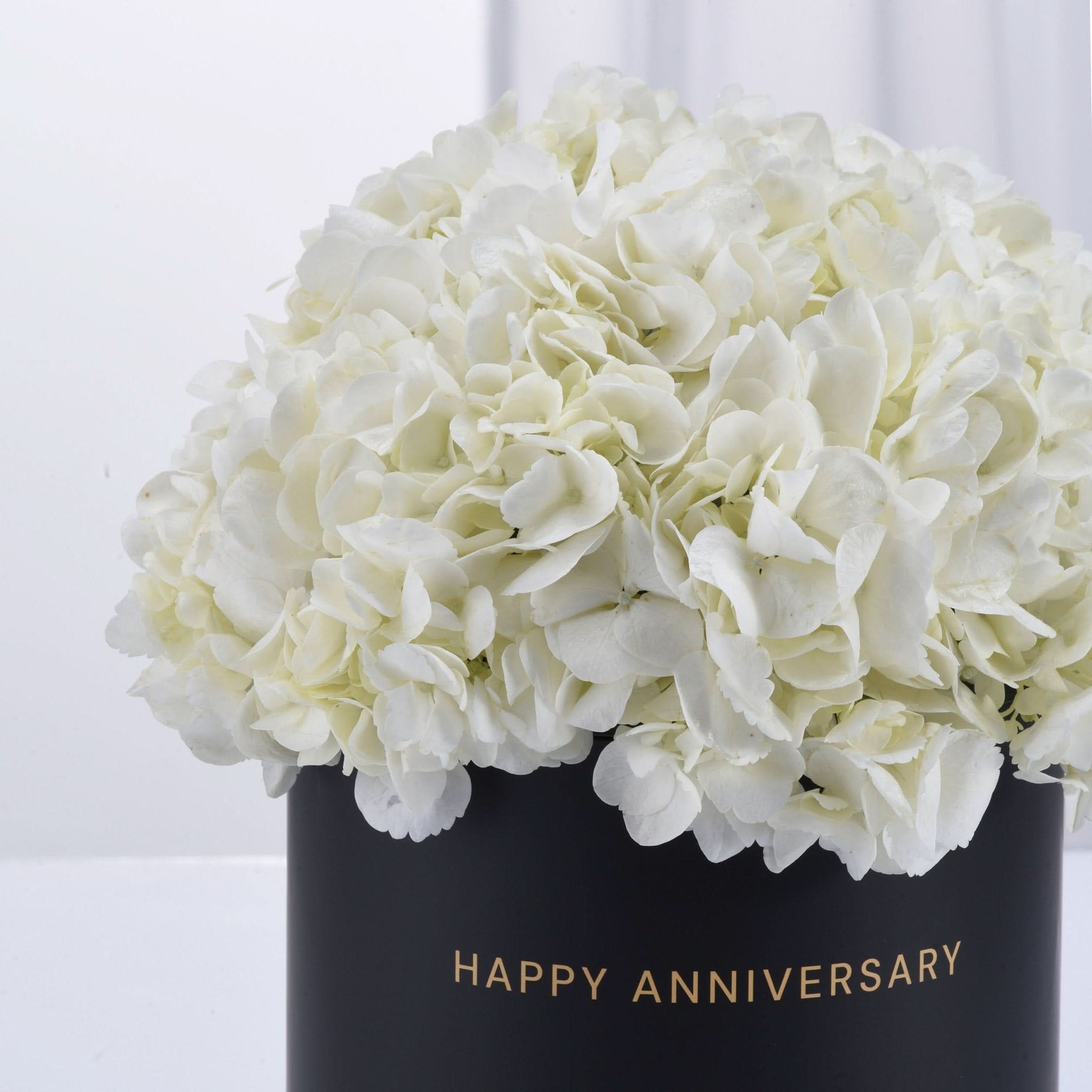 Luxurious white hydrangeas in matte black vase with gold "Happy Anniversary" text.