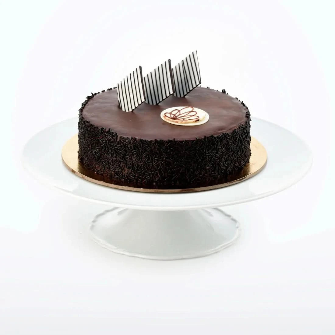 Rich chocolate cake adorned with striped chocolate shards and sprinkles on white stand.