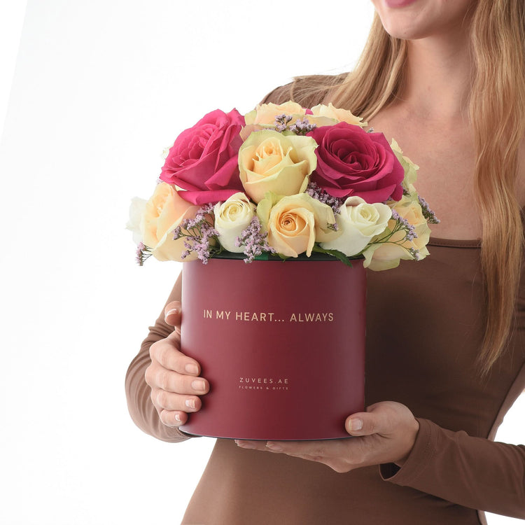 Rich burgundy box holds vibrant pink, yellow, and white roses with purple accents.