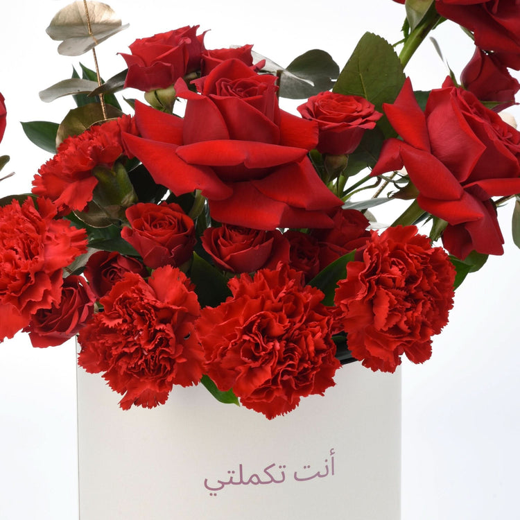 Vibrant red roses and carnations in white vase with Arabic script.