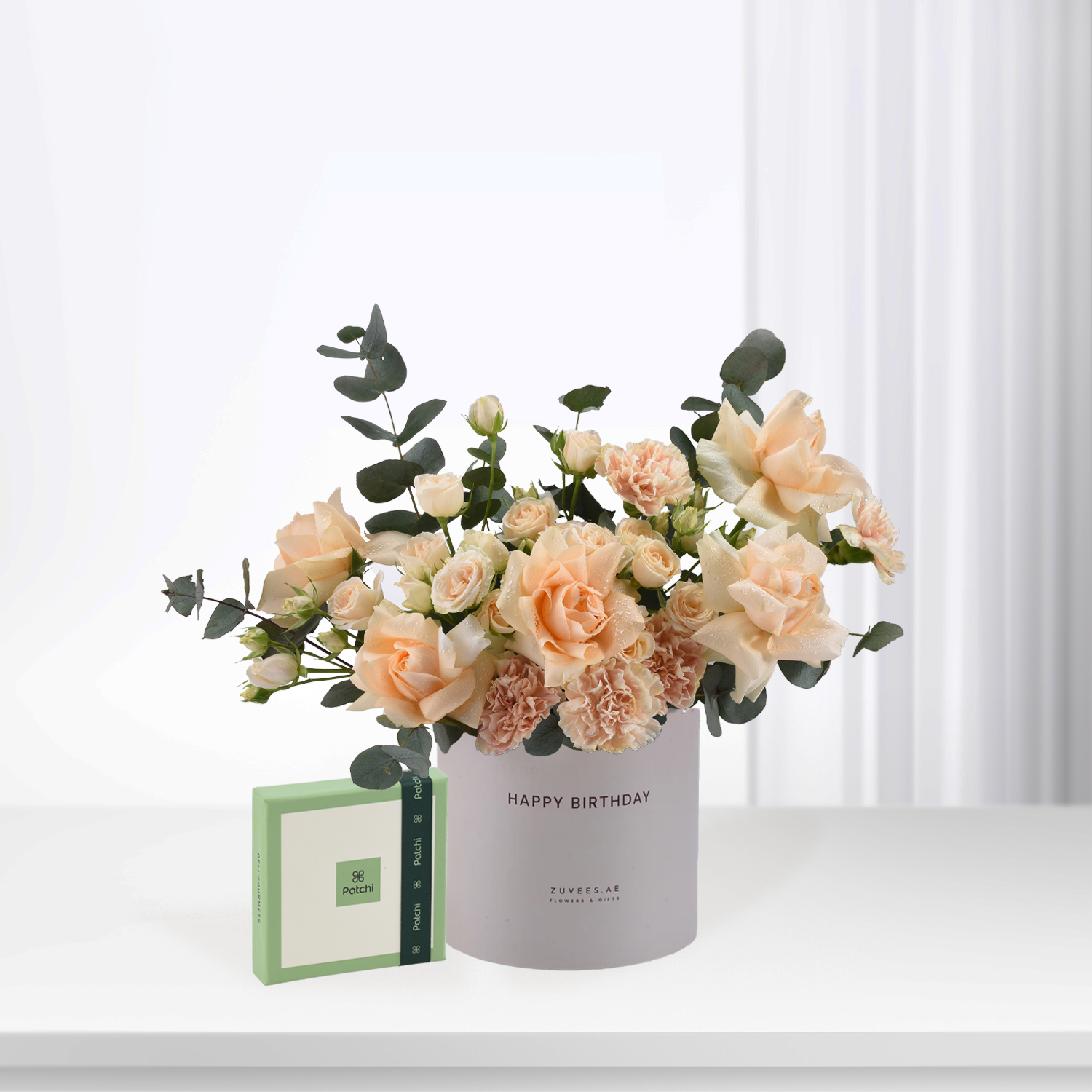 Soft peach roses and eucalyptus in white "Happy Birthday" vase beside green Patchi box.