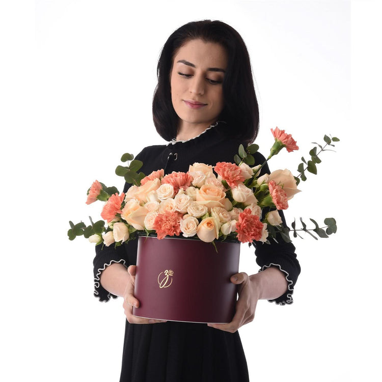 Woman holds burgundy hat box filled with peach roses and carnations.