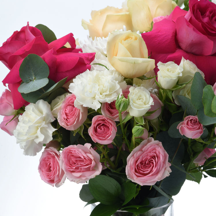 Vibrant pink, white, and yellow roses with lush greenery in clear vase.