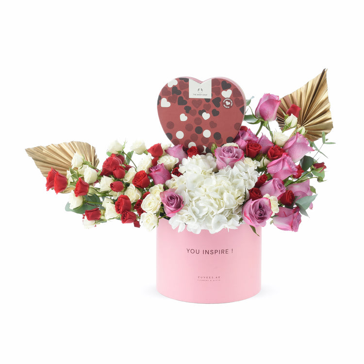 Pink cylinder vase with red, white, and pink roses, white hydrangeas, gold leaves, and heart-shaped chocolate box.
