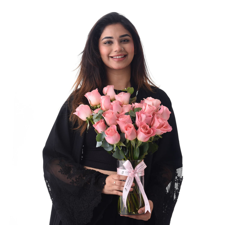 Smiling woman holds pink roses in clear vase with pink ribbon, wearing black lace robe.
