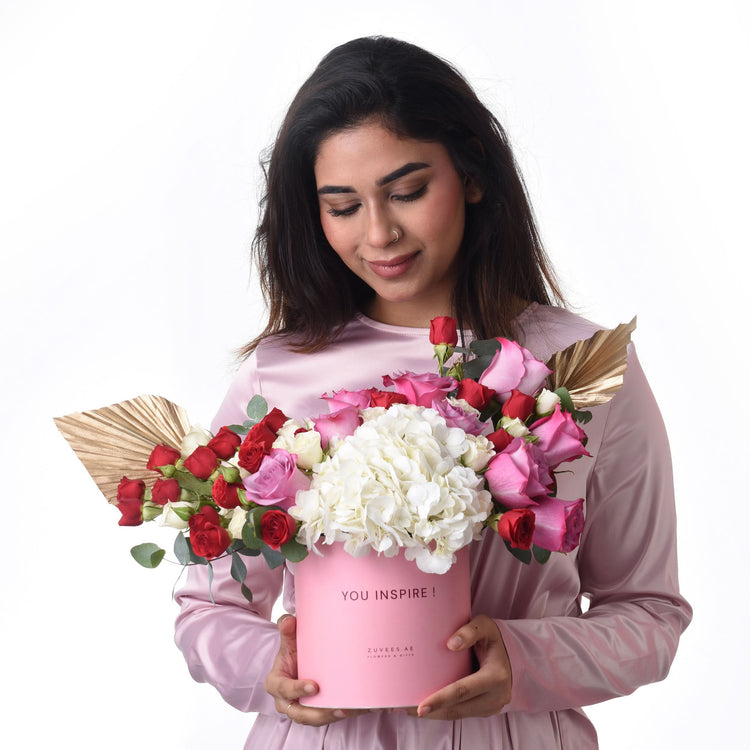 Pink box holds vibrant red, pink, white roses and hydrangeas, gold accents.