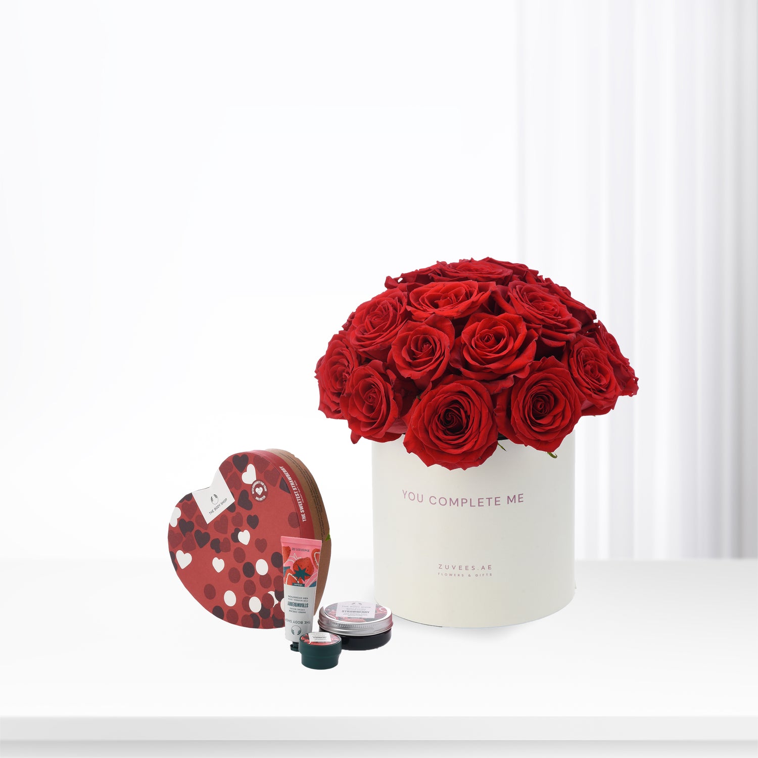 Vibrant red roses in white box with "You Complete Me" text, paired with heart-shaped strawberry-scented gift set.